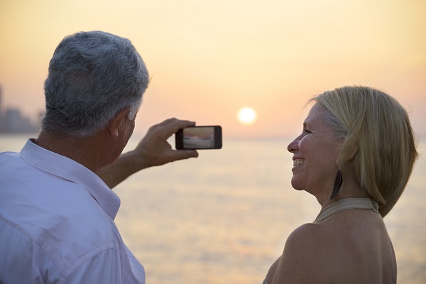 Senior man and woman using mobile phone to take photo on sunset over ocean