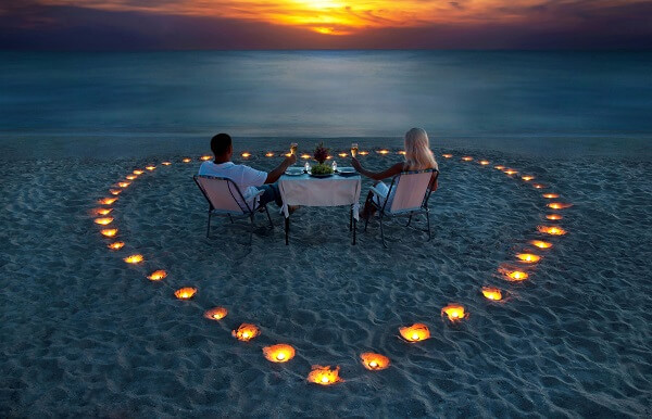 A young couple share a romantic dinner on the beach surrounded by candles in the shape of a love heart with sun setting in the background