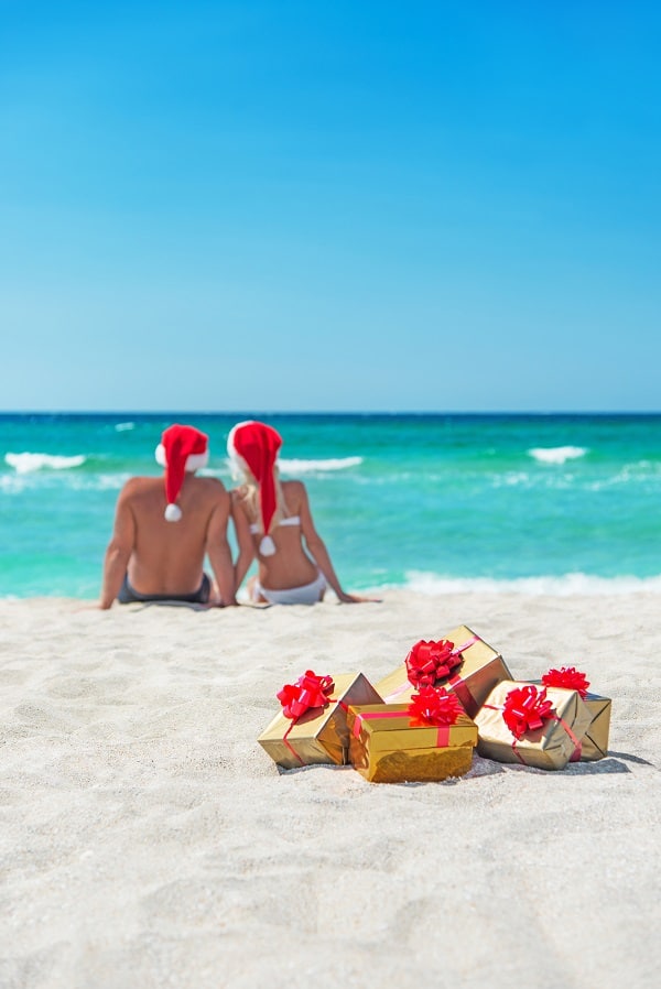 Couple sat on beach wearing Santa hats with wrapped golden Christmas presents behind them